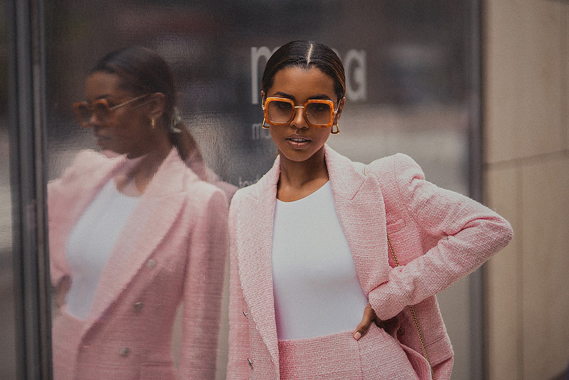 Selena Jutton adjusts her pink blazer beside a reflective storefront; her image mirrors back at her.