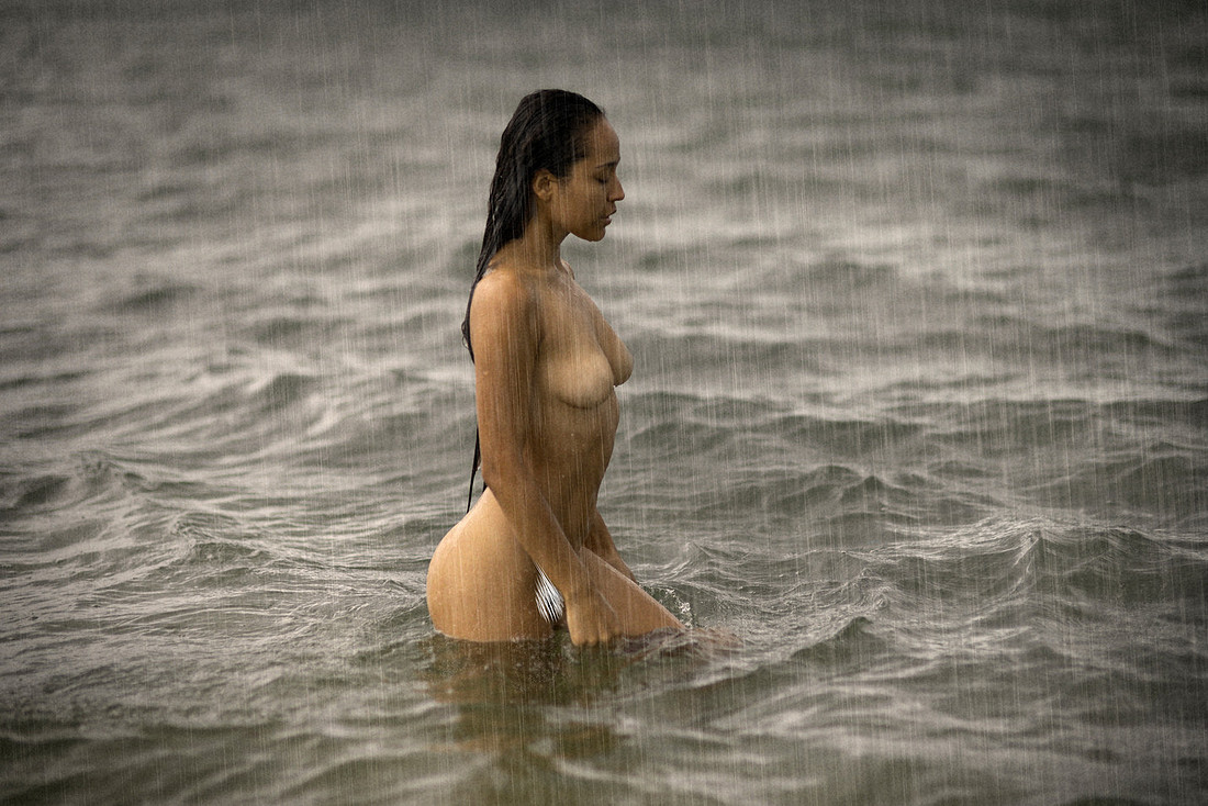 Model Becky Deeve stands, arms crossed over her chest, as rain streams down her glistening skin; water droplets highlight the curve of her shoulders and the strength in her stance.