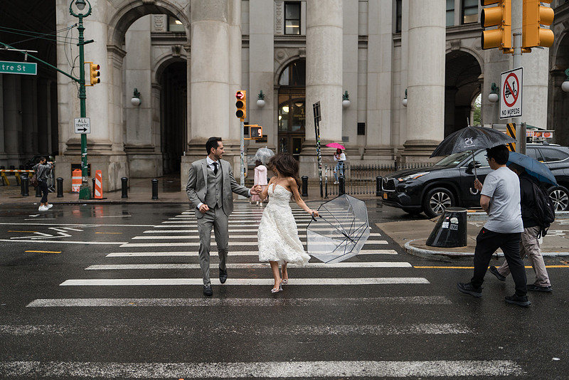 Bride and groom run across a rainy Centre Street crosswalk with a clear umbrella outside the Manhattan Municipal Building in NYC