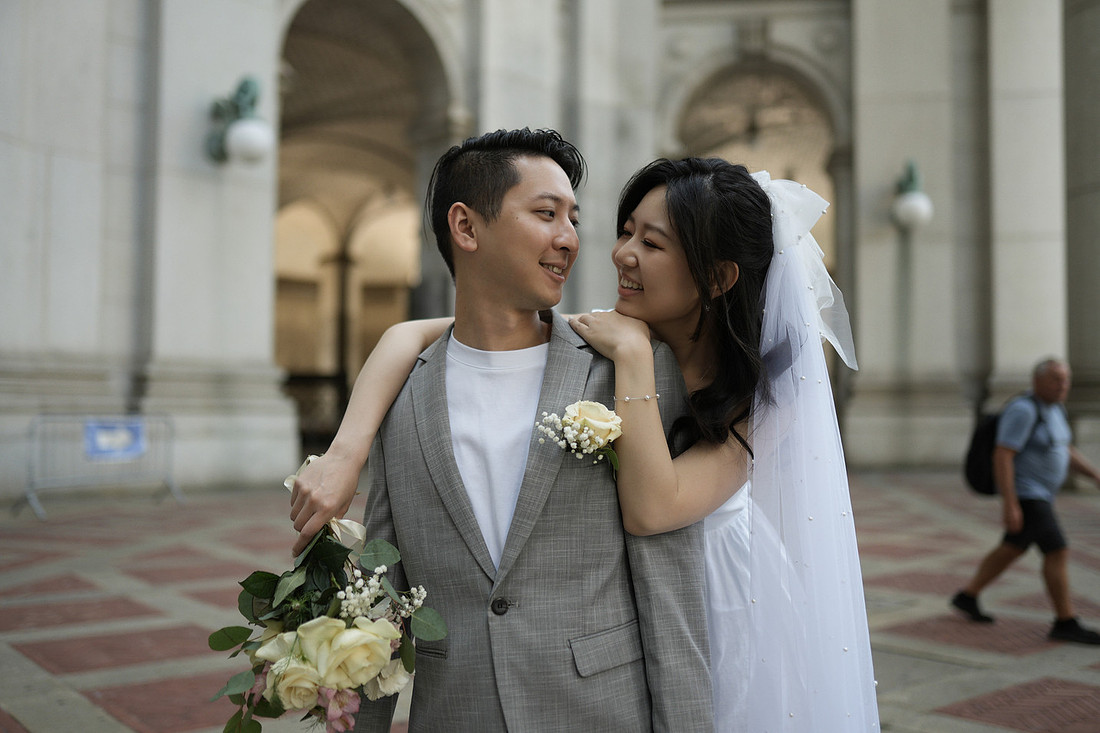 Newlywed couple smiling and embracing in front of an arched building after their City Hall wedding in New York City, with the groom holding a bouquet of flowers.